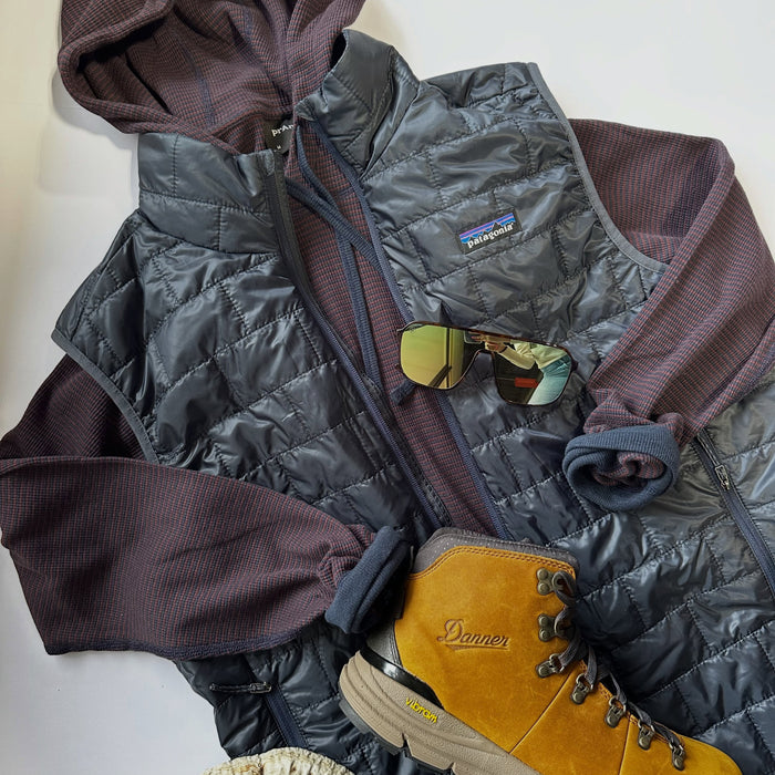 A styled flat-lay of outdoor apparel and accessories on a white background, featuring a dark gray quilted Patagonia vest over a brown ribbed hoodie, with a pair of sunglasses, a tan Patagonia hiking boot, and a rustic wood slice prop.